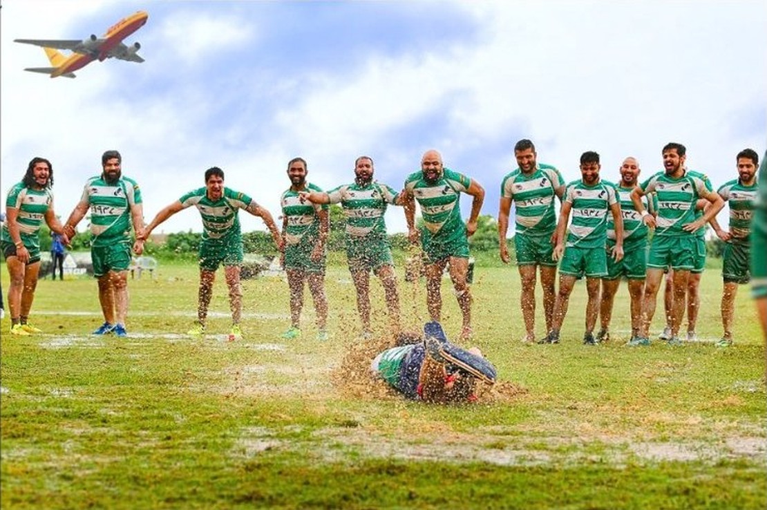 Team BOOMFA celebrating in custom Markhor kit — a player sliding through the grass as teammates erupt, with a plane flying overhead.
